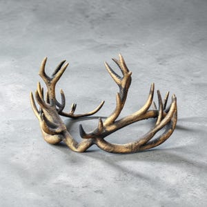 May include: A circular crown-shaped headpiece crafted to resemble a pair of deer antlers. The object features a textured, weathered gold and dark brown finish, with multiple branching points and curved tines extending upwards and outwards.