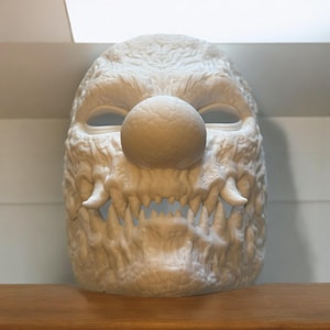 Peut inclure: Un masque en plastique blanc avec un grand nez rouge rond et un sourire narquois. Le masque a une surface texturée et semble être un monstre ou une créature.