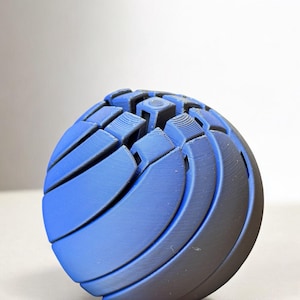 以下が含まれることがあります： 複雑な幾何学模様の青とグレーの球体。球体は、テクスチャのある表面を作り出す、湾曲した層状のセグメントで構成されています。色は青からグレーに変化し、デザインに深みを与えています。