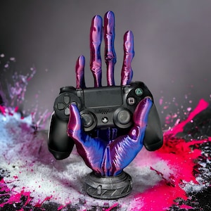 Könnte beinhalten: Ein schwarzer PlayStation-Controller, gehalten von einer 3D-gedruckten Hand mit einem blau-violetten, irisierenden Finish. Die Hand befindet sich auf einem schwarzen Sockel und der Hintergrund ist ein Spritzer aus weißer und rosa Farbe.