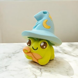 Op de afbeelding: Een grillig, geelgroen kikkerbeeldje met een lichtblauwe tovenaars hoed met gele maan- en steraccenten. De kikker heeft grote bruine ogen en een oranje mond, wat een charmant, fantasie-thema verzamelobject creëert.