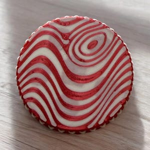May include: A round, red and white decorative ornament with a wavy, abstract design. The ornament has a white border.