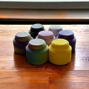 May include: A set of seven colourful bottle stoppers, including purple, green, yellow, and black. The stoppers are made of a smooth, rounded material.