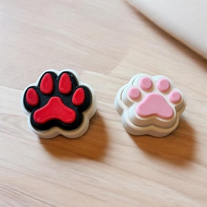 May include: Two paw-shaped cookie cutters. One has a black base with red paw pads, and the other is white with pink paw pads. Both have a white border and sit on a light wood surface.