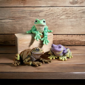 May include: Three frog figurines, one green, one brown, and one purple, sitting on a wooden surface. The green frog is sitting on a wooden block.