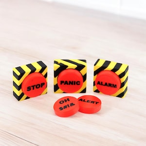 Op de afbeelding: Drie gele en zwarte vierkante knoppen met rode ronde knoppen waarop "STOP", "PANIC" en "ALARM" staat. Twee extra rode ronde knoppen zeggen "OH S#!&" en "ALERT".