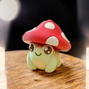 以下が含まれることがあります： 白い水玉模様の赤いキノコの帽子をかぶったカエルの気まぐれな置物。カエルは薄緑色で、お腹は黄色、目は大きく表情豊かです。木製の表面に置かれています。