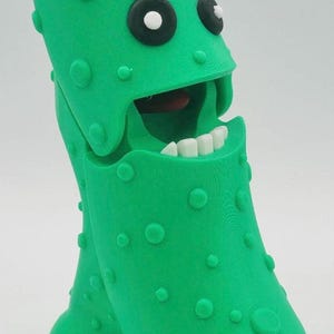 May include: A novelty figurine in the shape of a bright green pickle, featuring a wide open mouth with white teeth and a red interior. It has black and white eyes and raised circular bumps. A fun, quirky item.
