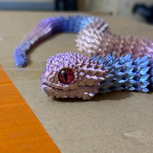 Op de afbeelding: Een regenboogkleurig, stekelig, slangachtig wezen met een rood oog. Het wezen is gemaakt van plastic en ligt op een bruin oppervlak.