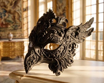 May include: A black, ornate masquerade mask with a bird-like design. The mask features intricate details, including swirling patterns and a wing-like extension. It is resting on a white surface, with a blurred background of a room with gold accents.