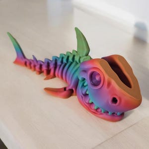 May include: A rainbow-colored plastic shark skeleton figurine with a hollow head. The figurine is made of multiple pieces that are connected together to form the skeleton.