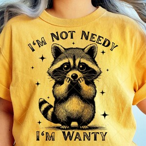 May include: Mustard yellow t-shirt featuring a detailed illustration of a raccoon with the text "I'M NOT NEEDY I'M WANTY". The raccoon is black and brown with a striped tail and is holding its paws up to its face. The shirt has a crew neck.