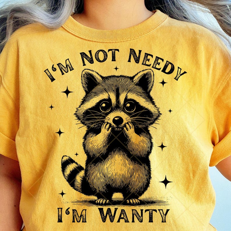 May include: Mustard yellow t-shirt featuring a detailed illustration of a raccoon with the text "I'M NOT NEEDY I'M WANTY". The raccoon is black and brown with a striped tail and is holding its paws up to its face. The shirt has a crew neck.