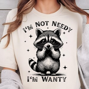 May include: Cream-colored t-shirt featuring a black and white illustration of a raccoon. The raccoon is depicted with large eyes and is covering its mouth with its paws. The text "I'M NOT NEEDY I'M WANTY" is printed above and below the raccoon.