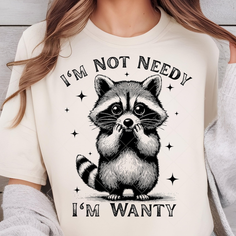May include: Cream-colored t-shirt featuring a black and white illustration of a raccoon. The raccoon is depicted with large eyes and is covering its mouth with its paws. The text "I'M NOT NEEDY I'M WANTY" is printed above and below the raccoon.