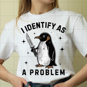May include: A white short-sleeve t-shirt featuring a graphic of a black and white penguin holding a knife. The text above the penguin reads I IDENTIFY AS and the text below the penguin reads A PROBLEM. The design includes small black star icons.