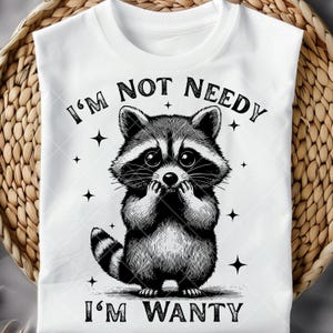 May include: White t-shirt featuring a black and white illustration of a raccoon with the text "I'M NOT NEEDY, I'M WANTY". The raccoon is drawn with its paws near its face, and the design includes small star accents.