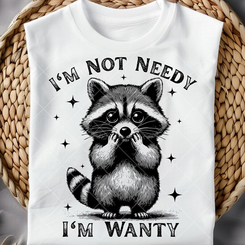May include: White t-shirt featuring a black and white illustration of a raccoon with the text "I'M NOT NEEDY, I'M WANTY". The raccoon is drawn with its paws near its face, and the design includes small star accents.
