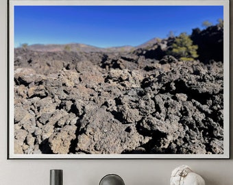 May include: A rectangular wall art print featuring a landscape of dark, jagged volcanic rock formations under a clear blue sky, displayed in a thin black frame above a dark wooden sideboard with a glass-topped desk and brown chairs.