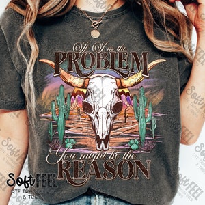 May include: A dark grey t-shirt featuring a graphic of a skull with horns in a desert landscape. The text "If I'm the Problem You might be the Reason" is printed on the shirt.