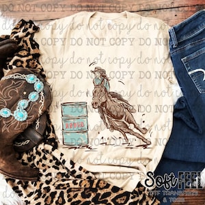 Könnte beinhalten: Ein beiges T-Shirt mit einem Aufdruck einer Frau, die auf einem Pferd um ein Fass reitet, auf dem "Rodeo" steht.