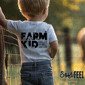 Pode incluir: Uma camiseta branca com texto preto que diz "Farm Kid" e "Raised on Dirt & Hard Work". A camiseta está sendo usada por uma criança pequena em pé na frente de uma cerca de madeira.
