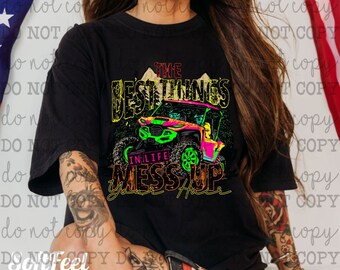 Peut inclure: T-shirt noir avec un graphique d'un véhicule tout-terrain rose et vert conduisant à travers un paysage montagneux. Le texte "The Best Things in Life Mess Up Your Hair" est imprimé sur le t-shirt.
