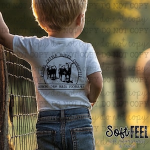 May include: A young child wearing a white t-shirt with a black and white graphic that says "Raising Cattle Builds Character, Working Them Builds Vocabulary". The child is standing in front of a metal fence.