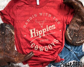 Op de afbeelding: Rood T-shirt met een wit grafisch ontwerp met de tekst "Hangin with the Hippies and the Cowboys". De grafiek bevat ook een schedel met hoorns.