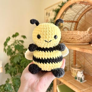 May include: A handmade crochet bee plushie. The bee features a yellow body with black stripes, black antennae, and black and white wings. The bee has a smiling face with black eyes. The plushie is a cute, soft toy.