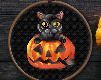 May include: A cross-stitch embroidery featuring a black cat with yellow eyes peeking out of an orange jack-o'-lantern. The embroidery is set in a wooden hoop against a dark wood background, with a pumpkin and succulent plant nearby.