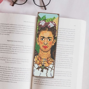 May include: A bookmark featuring a pixelated portrait of Frida Kahlo with flowers in her hair. The bookmark is made of fabric and has a brown wooden backing.