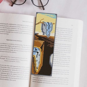 May include: A bookmark featuring a pixelated version of The Persistence of Memory by Salvador Dalí. The bookmark is made of fabric and has a blue, yellow, and brown color scheme.