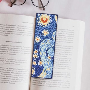 May include: A blue and yellow bookmark featuring a pixelated image of a starry night sky. The bookmark is made of fabric and has a fringed edge. The bookmark is lying on top of an open book.