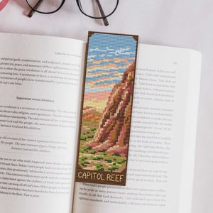 May include: A bookmark featuring a cross-stitch style image of Capitol Reef National Park in Utah. The bookmark is made of wood and has a brown frame. The image shows a red rock formation with a blue sky and white clouds in the background. The text "Capitol Reef" is printed in white on the bookmark.