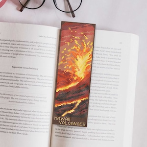 May include: A handmade bookmark featuring a pixelated design of a volcano with orange, yellow, and brown hues. The text "HAWAII VOLCANOES" is at the bottom. The bookmark is placed in an open book.