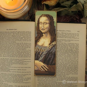 May include: A close-up of a book with a bookmark featuring a beaded portrait of the Mona Lisa. The bookmark is on a book titled "The Pickwick Club" and "The Consequence of  Fate". A lit candle and a cup of tea are in the background.