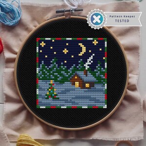 May include: A completed cross-stitch embroidery in a black hoop. The design features a winter night scene with a house, trees, stars, and a crescent moon. The border is red and green. The text "Pattern Keeper TESTED" is visible.