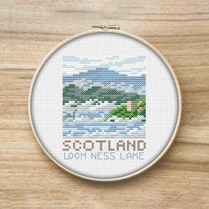 May include: A cross-stitch embroidery of Loch Ness Lake in Scotland, presented in a wooden hoop. The design depicts a landscape with blue water, green hills, and a castle, under a blue sky. The text "SCOTLAND LOCH NESS LAKE" is stitched below.