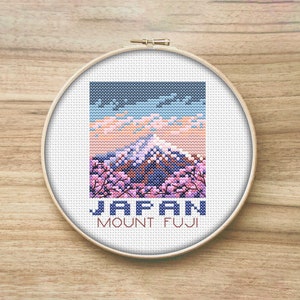 以下が含まれることがあります： 日本の富士山をクロスステッチで刺繍した白い刺繍枠。デザインは、ピンクとブルーの空に雪を頂いた山とピンクの桜の花が描かれています。
