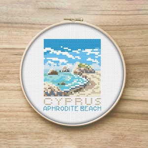 May include: A cross-stitch embroidery hoop with a white background and a colorful image of a beach scene with rocks, water, and sand. The text "CYPRUS APHRODITE BEACH" is printed below the image.