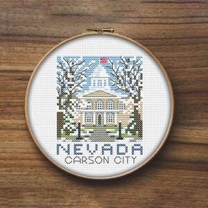 May include: Cross-stitch embroidery of the Nevada State Capitol building in Carson City, Nevada. The building is white with a green roof and a small American flag on top. The building is surrounded by trees and snow. The text "NEVADA CARSON CITY" is below the building.