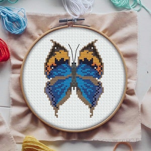 May include: A cross-stitch butterfly design in a wooden embroidery hoop. The butterfly has blue, orange, and brown wings. The embroidery is on beige fabric, with colourful embroidery threads around it.