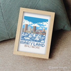 May include: A framed cross-stitch design featuring a cityscape of Baltimore, Maryland. The design includes a blue sky with white clouds, a skyline of buildings, and boats in the water. The text "MARYLAND BALTIMORE" is stitched below the cityscape.