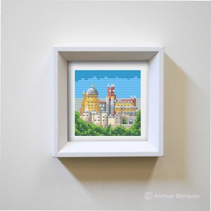 May include: A framed cross-stitch embroidery depicting a yellow and red building with a blue sky and green trees in the foreground.