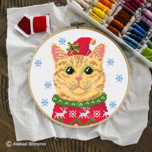 May include: Cross-stitch embroidery of a ginger cat wearing a red Christmas hat and sweater with snowflakes. The cat has green eyes and a white muzzle. The embroidery is in a wooden hoop, with spools of thread in the background.