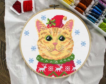 Könnte beinhalten: Kreuzstich-Stickerei einer rot getigerten Katze mit rotem Weihnachtshut und Pullover mit Schneeflocken. Die Katze hat grüne Augen und eine weiße Schnauze. Die Stickerei befindet sich in einem Holzreifen, mit Garnrollen im Hintergrund.