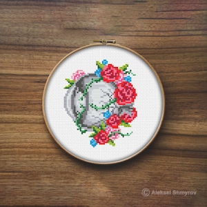 May include: A white embroidery hoop with a cross-stitch design of a letter D surrounded by red, pink, and blue flowers.