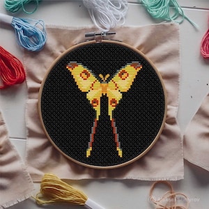 Puede incluir: Un aro de bordado de punto de cruz con fondo negro y una mariposa amarilla con detalles rojos y grises.