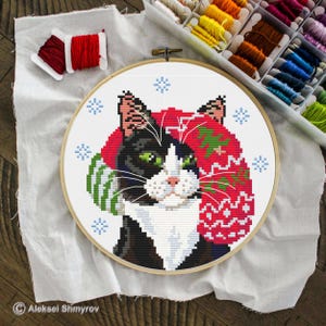 Peut inclure: Un cercle de broderie au point de croix représentant un chat noir et blanc portant un bonnet et une écharpe d'hiver rouges et verts. Le chat a les yeux verts et est entouré de flocons de neige bleus. Du fil à broder et du fil sont visibles en arrière-plan.